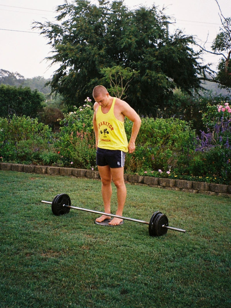 Person in a yellow Barbell Row stringer standing over a barbell on a garden lawn